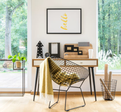 Blanket on designer chair at desk in bright work area with poster, plant and view on a forest
